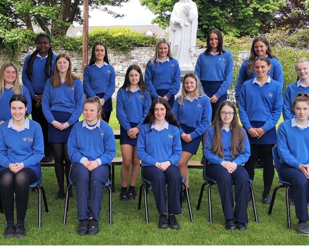 Un gruppo di ragazze irlandesi su tre file posa per la foto di classe in un giardino; indossano una divisa scolastica blu