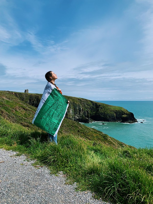 Da una scogliera, una ragazza avvolta in una bandiera irlandese guarda il cielo e il mare davanti a lei; sullo sfondo scogliere a picco sull’oceano