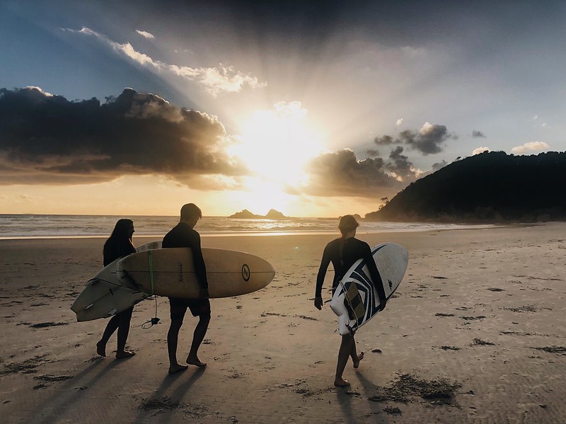 Trois jeunes marchent de dos sur une plage au coucher du soleil avec une planche de surf sous le bras.