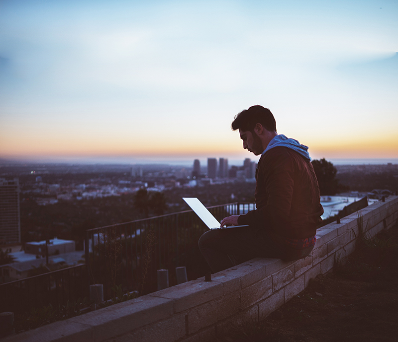 Un jeune homme est assis sur un muret au coucher du soleil avec un ordinateur portable sur les genoux.