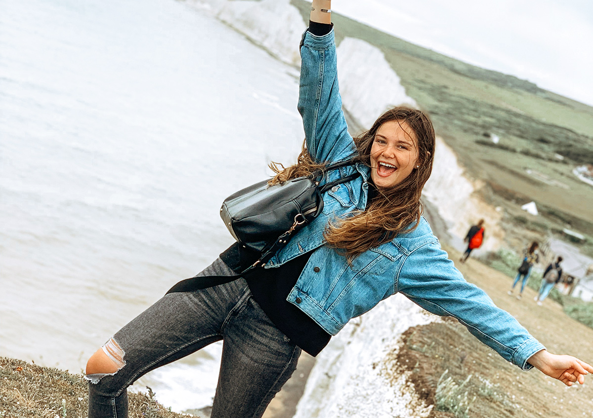 Une jeune fille pose souriante, les bras levés, devant un paysage de falaises de la côte de l'Angleterre.