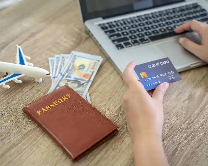 Su un tavolo ci sono un passaporto, del denaro in contanti, un piccolo aereo, un pc e una mano con una carta di credito