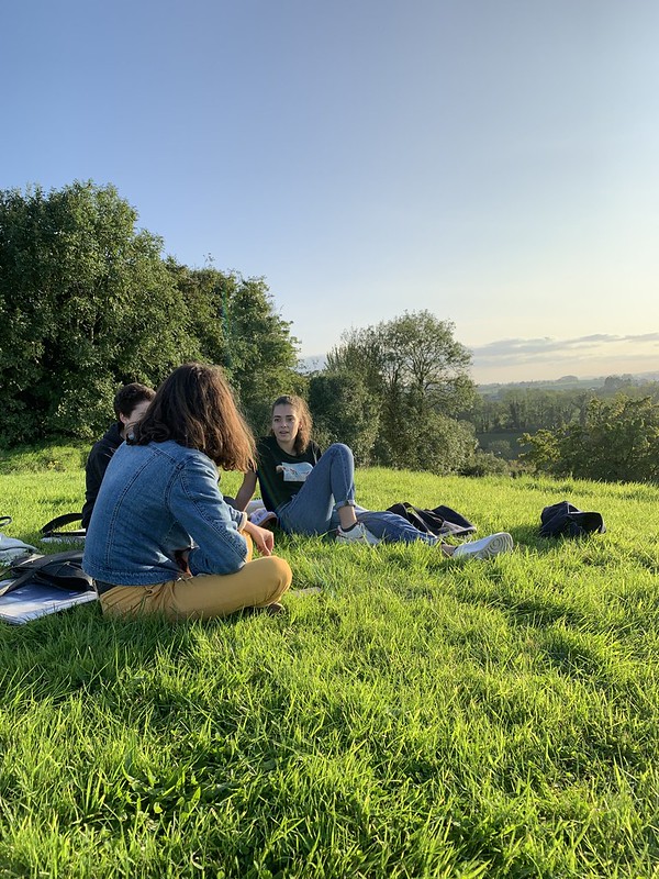 Alcune ragazze irlandesi in abiti sportivi sono sedute rilassate su un campo verdeggiante, cielo azzurro sullo sfondo