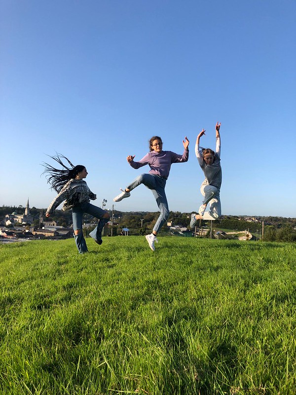Tre studentesse irlandesi, sorridenti e allegre, spiccano un salto su un prato verde chiaro; cielo azzurro dietro di loro