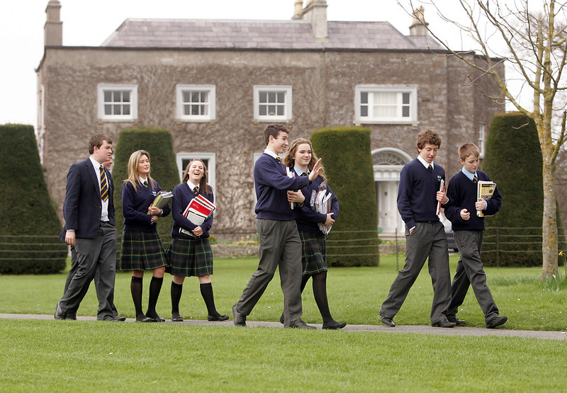 Un gruppo di studenti e studentesse irlandesi con indosso la divisa passeggiano nel cortile erboso di una scuola, che compare sullo sfondo