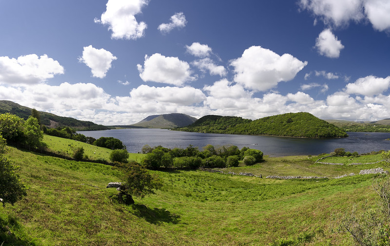 Un tipico paesaggio irlandese un prato scosceso verde brillante, alcuni alberi dalle folte chiome, un lago azzurro e un cielo a pecorelle