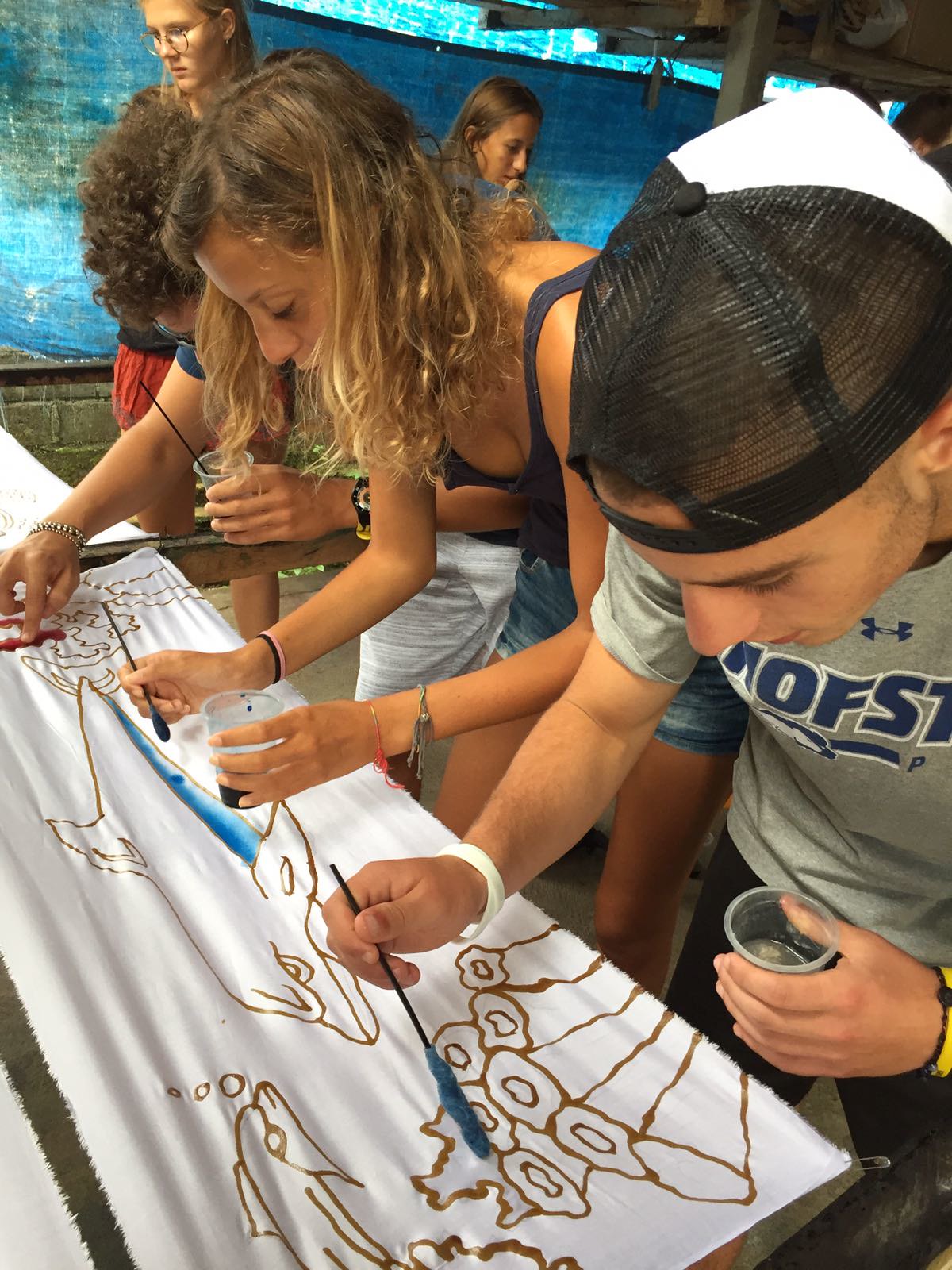 Tre giovani in abiti estivi sono chini su un lenzuolo bianco: stanno colorando con gli acquerelli le sagome di due delfini, alghe e coralli