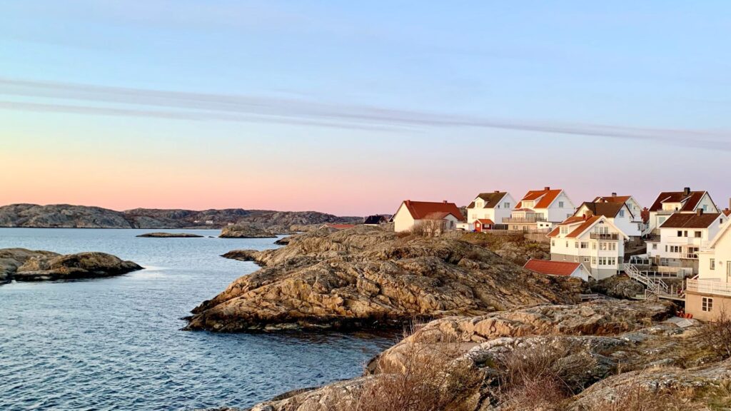 Natura svedese all’alba: terra rocciosa e brulla che entra in un calmo mare azzurro chiaro. Tipiche case nordiche in legno bianche con tetti rossi