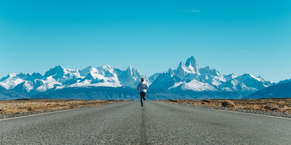 Ragazzo adolescente in jeans e felpa chiara, di spalle corre su una strada asfaltata verso le punte innevate delle montagne argentine