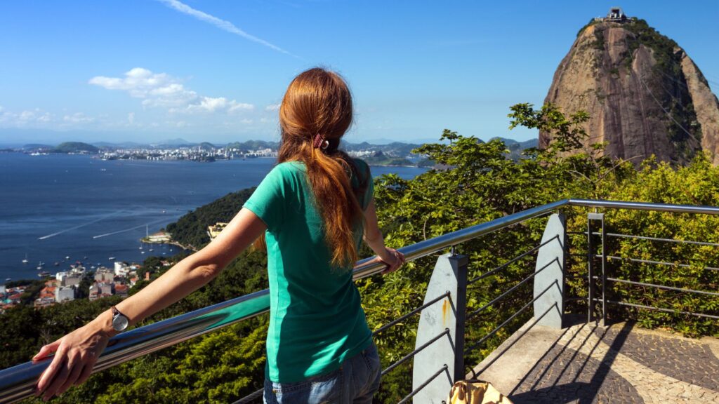 Ragazza di spalle ammira una vista dall'alto di una costa brasiliana, davanti a lei c'è il mare, una foresta e una montagna stretta e alta
