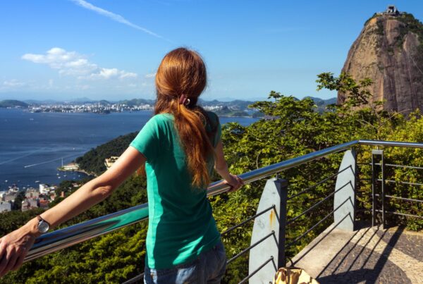 Ragazza di spalle ammira una vista dall'alto di una costa brasiliana, davanti a lei c'è il mare, una foresta e una montagna stretta e alta