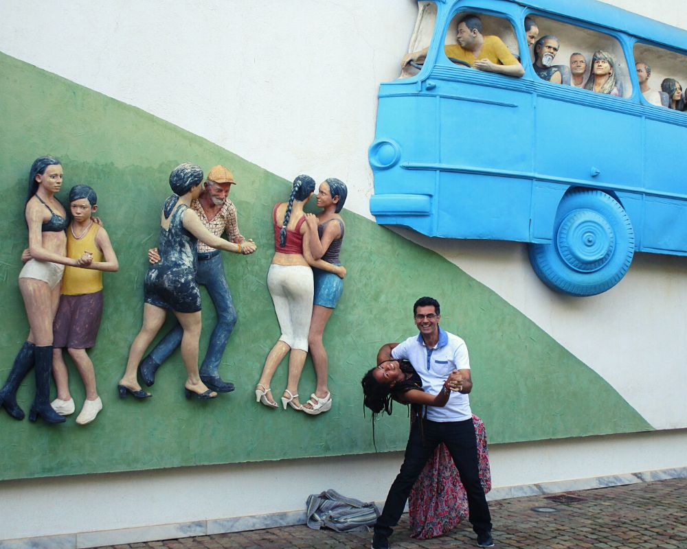 Un ragazzo e una ragazza ballano davanti a un murales che celebra la danza in brasile, sul muro c'è anche un furgoncino azzurro