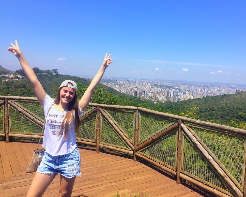 Ragazza con braccia alzate sorride alla fotocamera, alle sue spalle un panorama di una foresta brasiliana e in lontananza una città