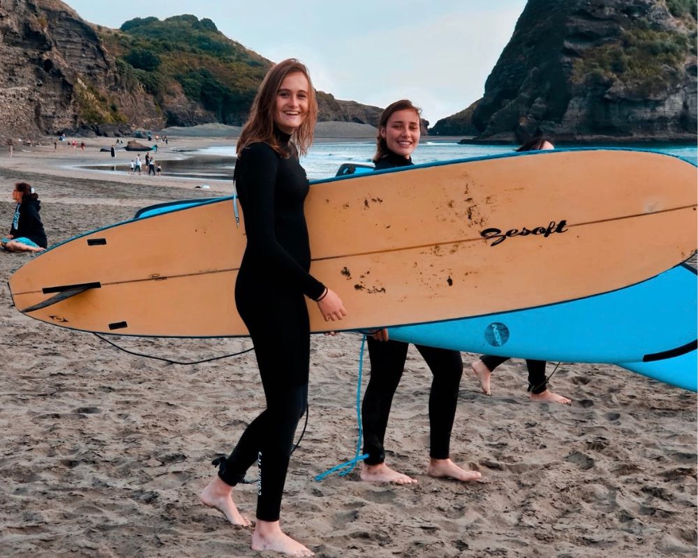 Una giovane con capelli lunghi biondi e una con capelli castani raccolti sorridono con due tavole da surf in mano; sullo sfondo terre a picco sul mare e acqua