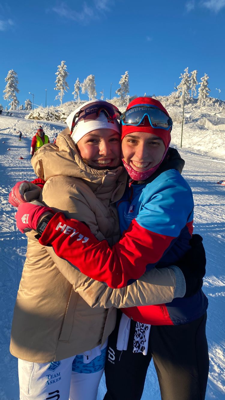 Sulla sx ragazza norvegese con tuta da sci bianca e giacca beige; sulla dx studente di scambio con pantaloni neri e giacca rossa e azzurra; si abbracciano sulle piste da sci