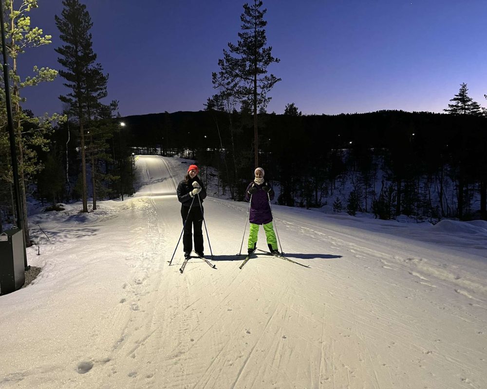 Un’ampia pista da fondo illuminata e costeggiata da conifere scure, un cielo serale e due ragazze con sci nei piedi in posa di fronte alla fotocamera