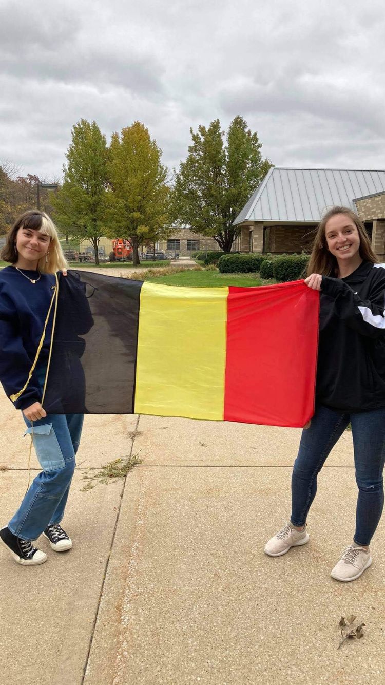 Nel cortile di casa, una ragazza con capelli a caschetto metà neri e metà biondi e una ragazza con capelli lunghi castani tengono una bandiera belga