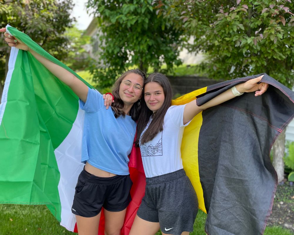 Due ragazze abbracciate e sorridenti in abiti estivi tengono dietro di loro una bandiera italiana e una belga; sullo sfondo giardino con alberi