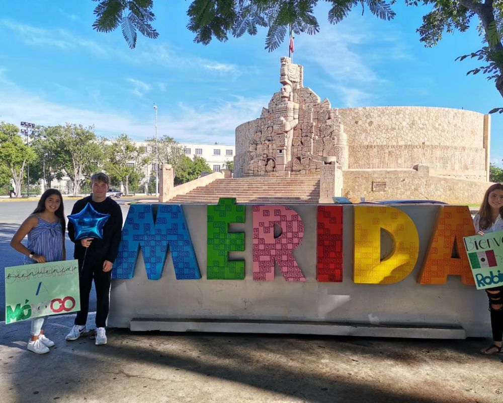 Una ragazza messicana e un exchange student davanti alla scritta colorata di benvenuto a Merida, in Messico; alle spalle un monumento della città