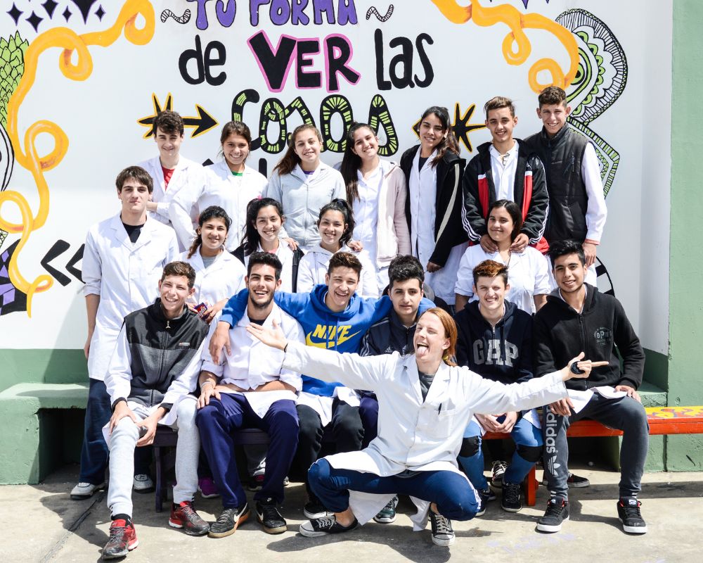 Alcuni ragazzi argentini che indossano un camice bianco sono disposti su tre file e sorridono alla fotocamera. Sullo sfondo un muro bianco con scritte colorate