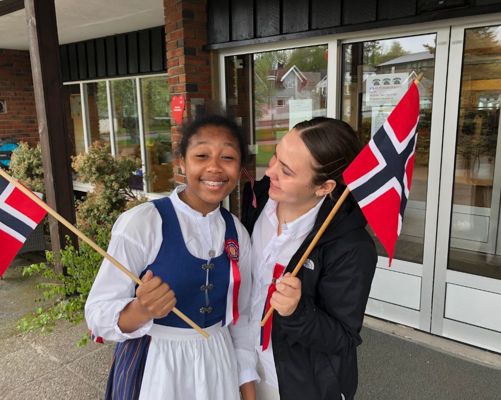 Una ragazza di colore e una ragazza bianca con una bandierina norvegese in mano ed abbigliamento tipico. Dietro, vetrate di una scuola in legno e mattoni