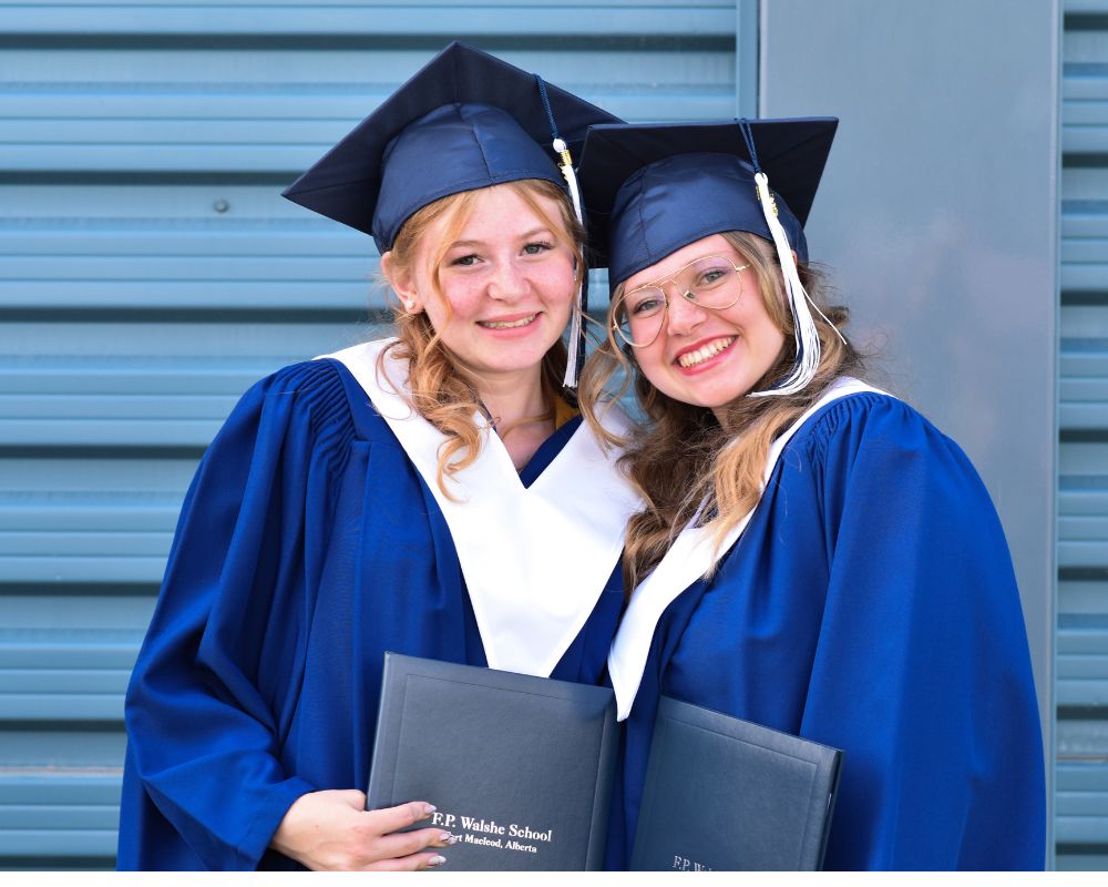 Due ragazze bionde sorridono alla fotocamera. Indossano una toga blu con colletto bianco e unl tocco blu. In mano tengono il diploma rilasciato dalla scuola