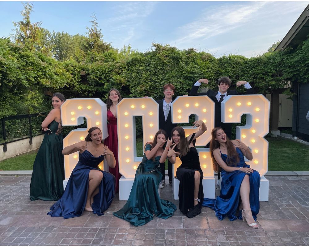 Due ragazze e sei ragazzi in abiti eleganti posano intorno a una scritta luminosa che riporta la scritta 2023 in occasione del ballo di fine anno