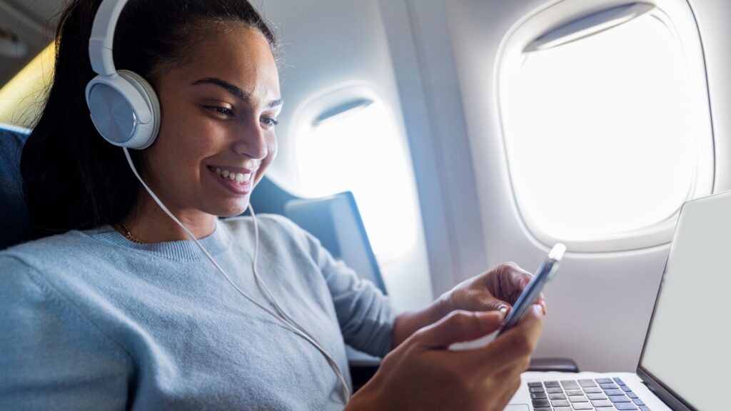 Cosa fare in aereo: ragazza con cuffie, cellulare e computer portatile seduta vicino al finestrino