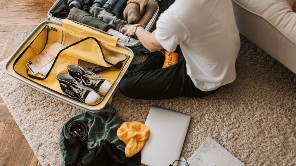 Foto di una valigia gialla aperta con scarpe, vestiti, portatile a terra e uomo di spalle che inserisce maglioni all'interno