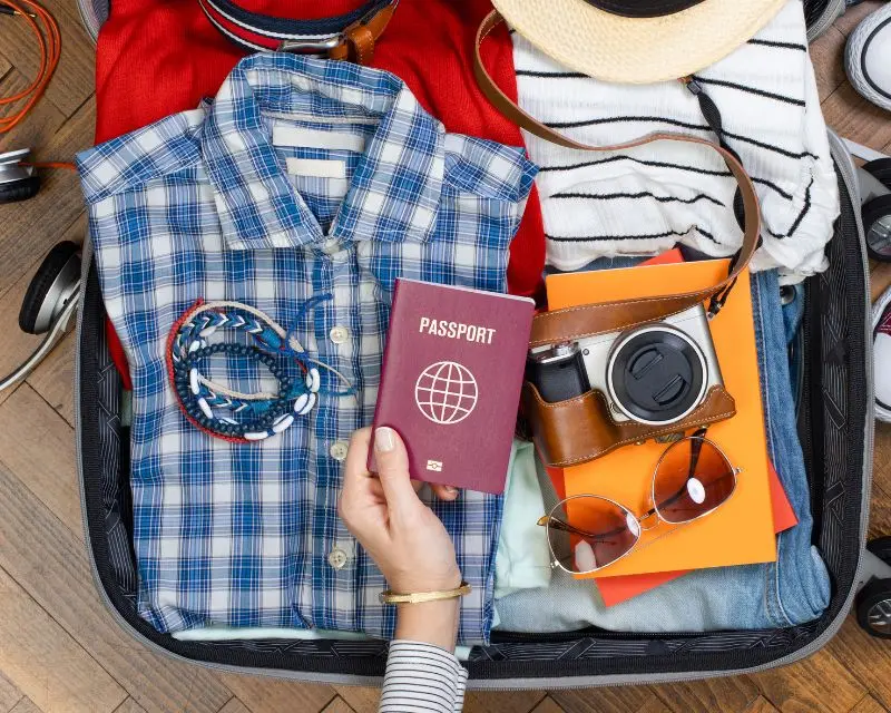 Una valigia con camicia blu a quadretti, magliette a righe, jeasn, occhiali dal sole, braccialetti, quaderni, macchina foto e passaporto