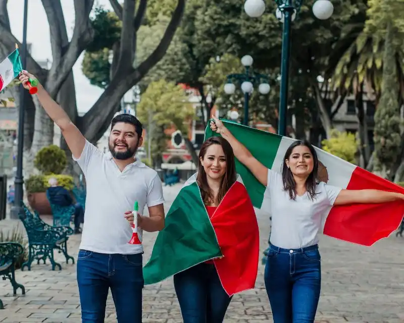 Un uomo e due donne in strada con trombette e bandiere del Messico. Indossano jeans e maglietta bianca