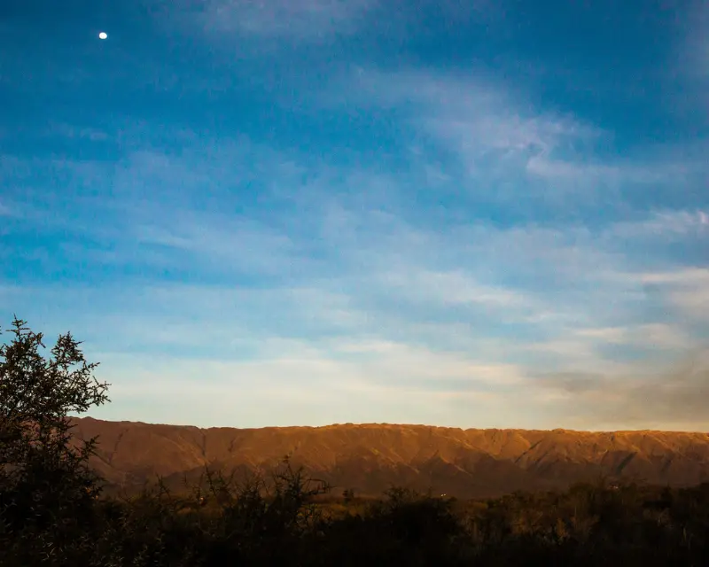 Panorama con montagne dell'Argentina e alberi in primo piano, cielo azzurro e la luna che fa capolino