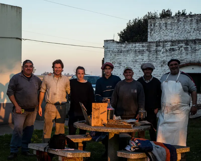 Gruppo di uomini in primo piano che sorridono in occasione di una grigliata con la tipica carne asado. Lo spazio intorno è semplice con una calda luce del tramonto