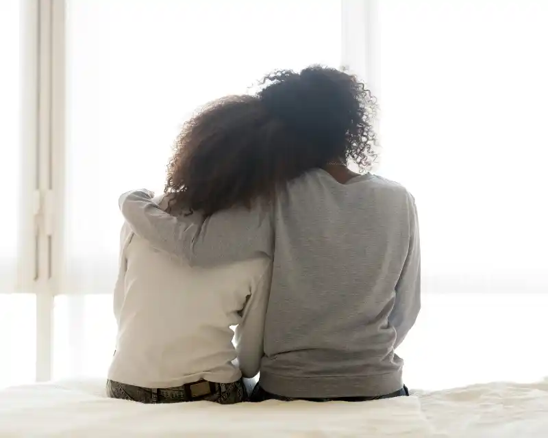 Due persone sono sedute su un letto e si abbracciano: un momento di vicinanza che esprime il legame profondo tra genitori e un figlio lontano da casa.
