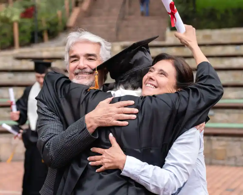 Un ragazzo con toga e diploma abbraccia i genitori sorridenti: un momento di gioia che racconta l’orgoglio, la distanza di un figlio lontano da casa e la crescita insieme.