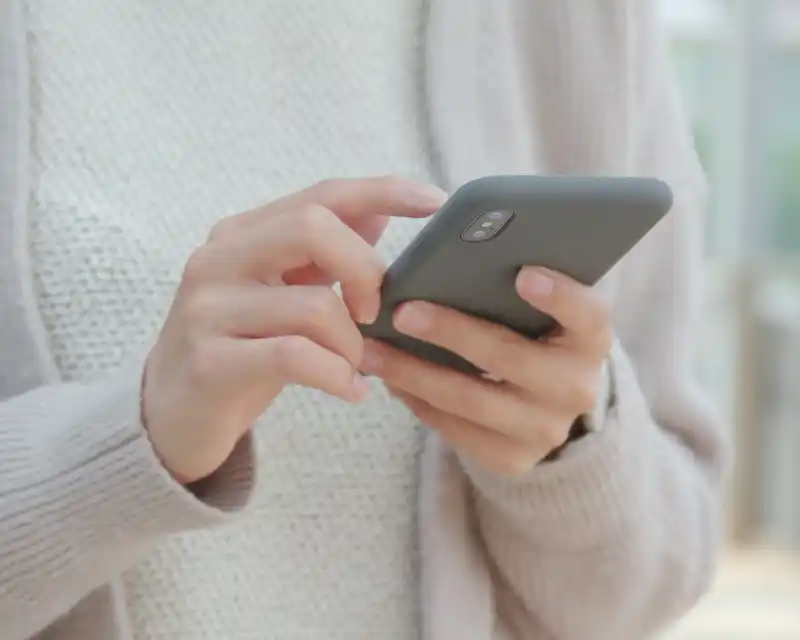 Primo piano delle mani di un adulto che tiene e usa uno smartphone: un gesto quotidiano che racconta il bisogno di contatto con un figlio lontano da casa.