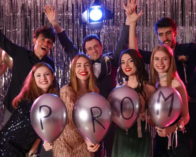 Gruppo di ragazzi e ragazze in vestiti da ballo di fine anno sorreggono palloncini con scritta Prom, tipici dei balli di fine anno USA.