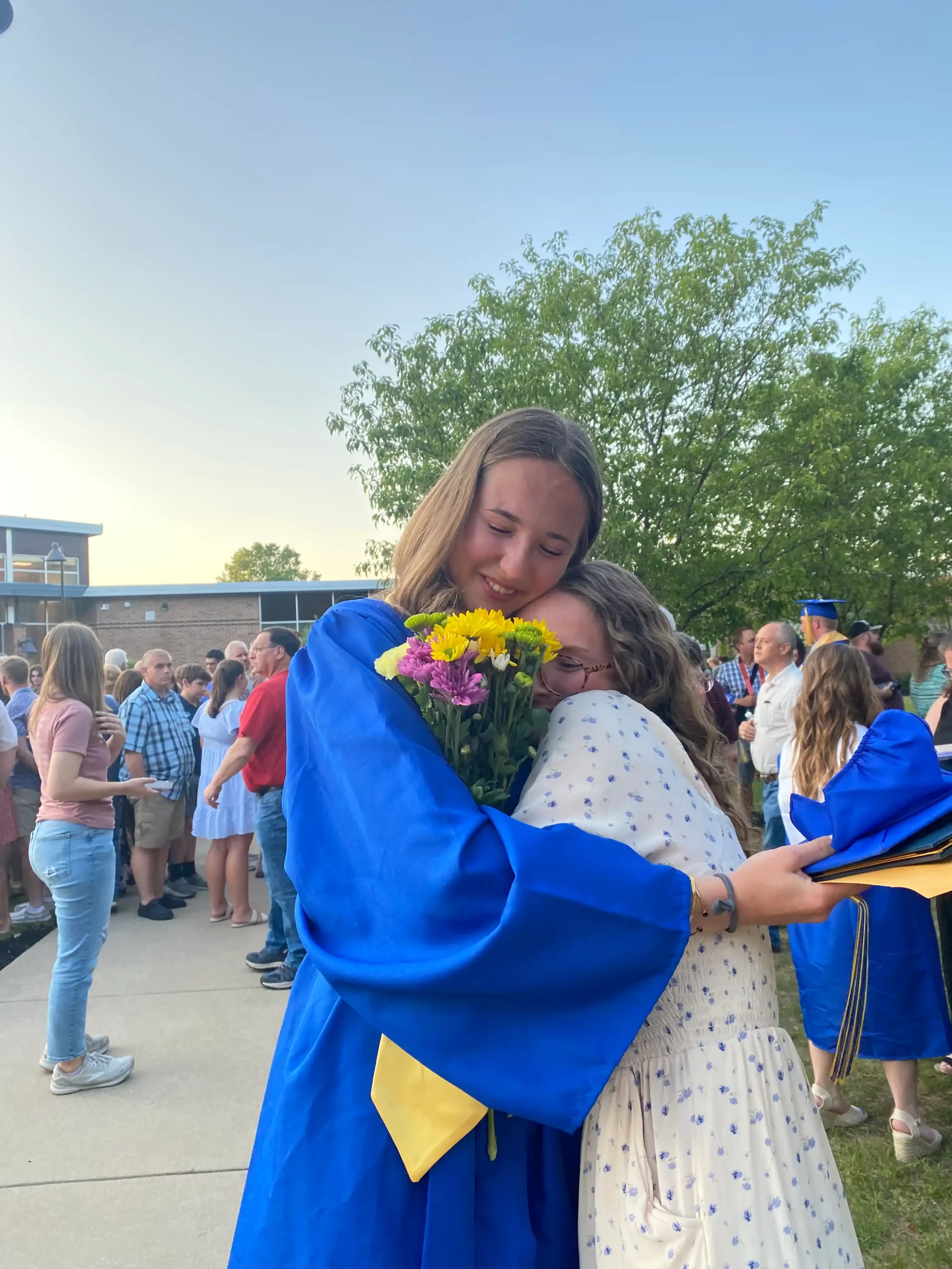 Ragazza in toga blu abbraccia un’amica con fiori in mano durante la cerimonia di diploma americano.