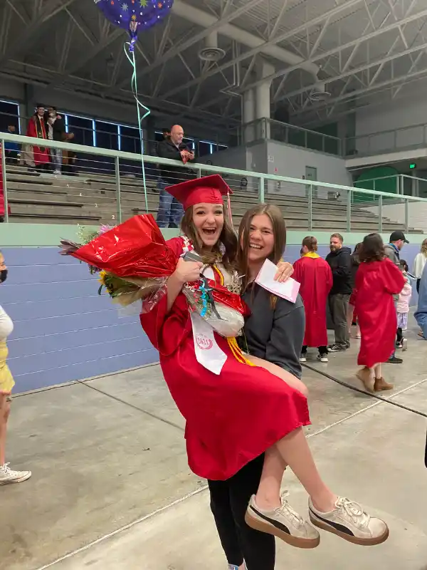 Studentessa in toga rossa viene sollevata in braccio da un’amica mentre festeggia il graduation day con entusiasmo e fiori.
