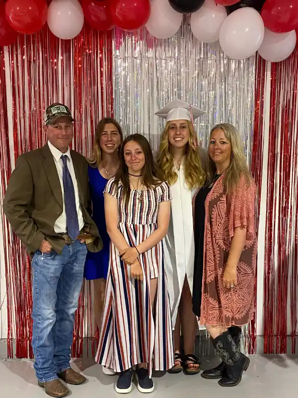 Ragazza in toga bianca posa con la sua famiglia ospitante davanti a decorazioni per celebrare la graduation negli USA.