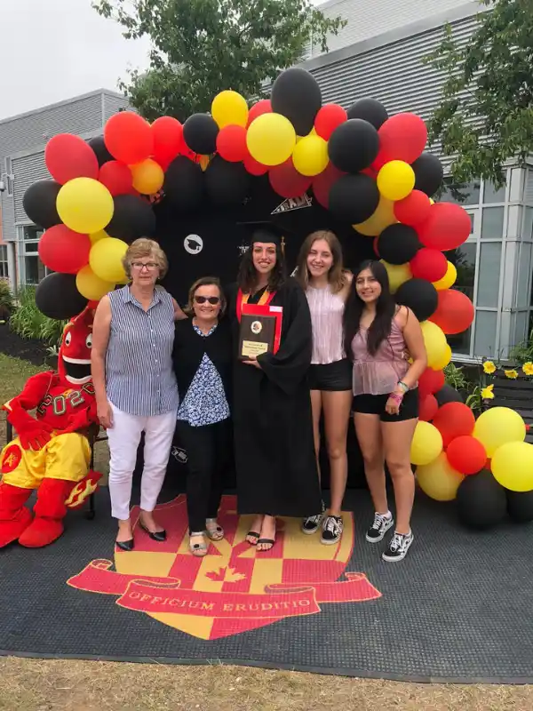 Ragazza in toga rossa festeggia la sua graduation americana con la famiglia ospitante sotto un arco di palloncini colorati.