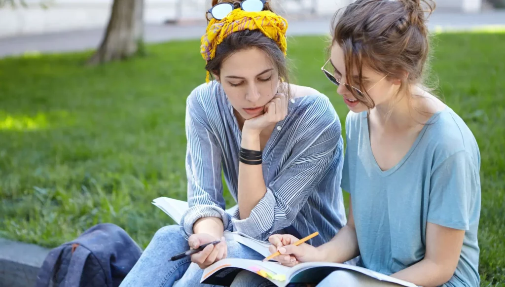 Ragazze sedute vicino a un prato fanno degli esercizi di grammatica su un libro di testo per imparare il tedesco
