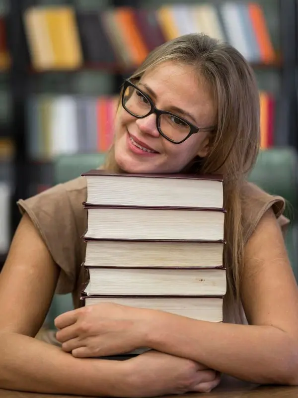 Ragazza con gli occhiali e i capelli biondi stringe tra le braccia sorridente una pila di libri per imparare il tedesco da autodidatta
