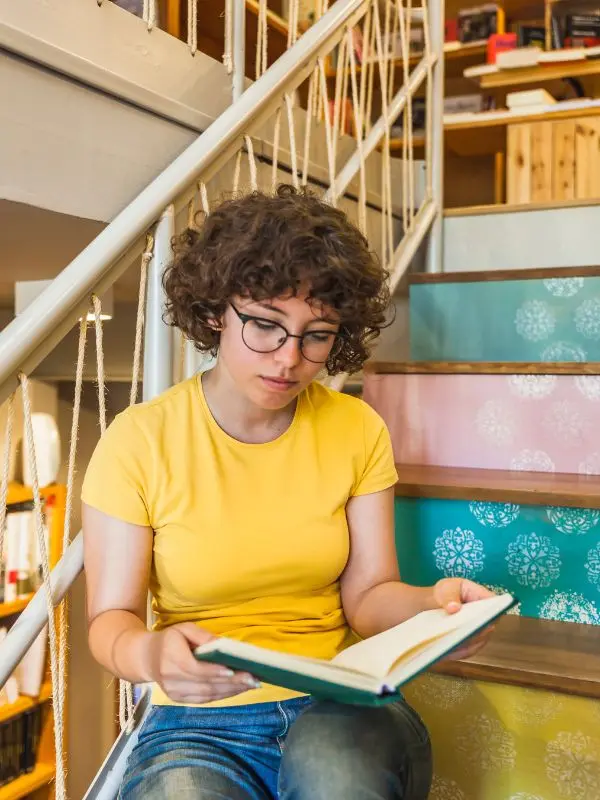 Ragazza con maglietta gialla, occhiali e capelli ricci legge sulle scale un libro per imparare il tedesco