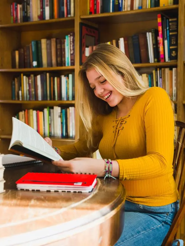 Una giovane ragazza felice con maglietta gialla e capelli biondi legge in biblioteca gli appunti presi dal suo libro per imparare il tedesco
