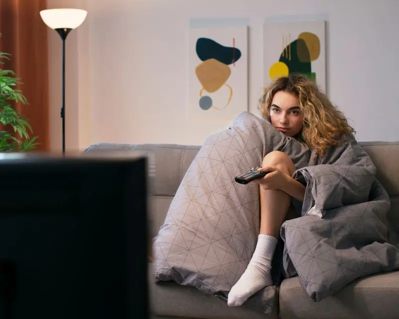 Ragazza bionda infreddolita sul divano con una coperta grigia addosso guarda una serie TV in tedesco