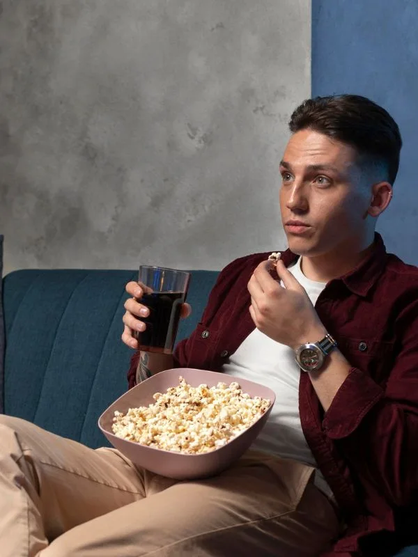 Ragazzo quindicenne con una camicia bordeaux, beve una coca e mangia pop corn mentre guarda una serie TV in tedesco