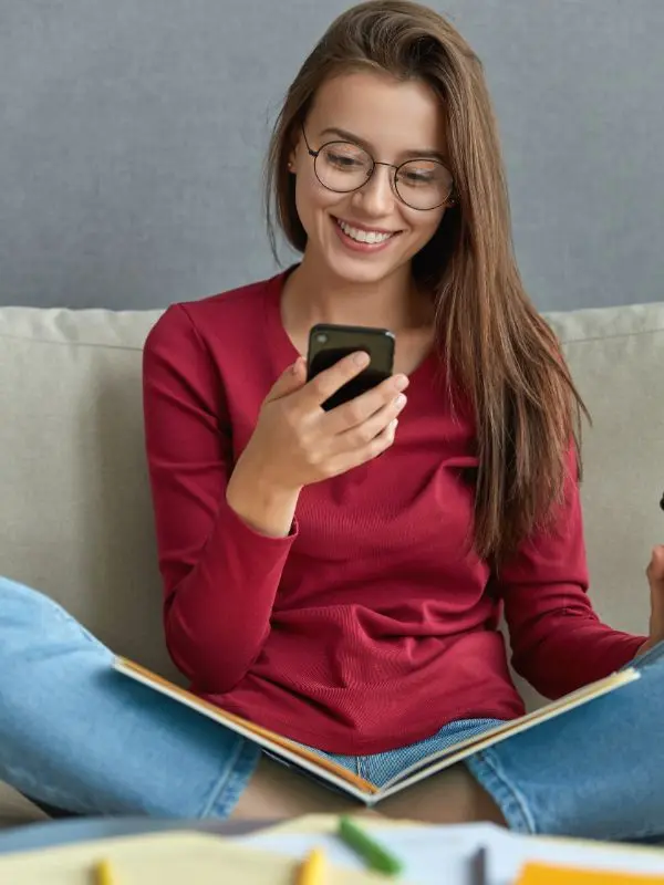 Ragazza con i capelli lisci lunghi e una maglietta rossa sta seduta sul divano, mentre studia tedesco con l'utilizzo di un'applicazione.