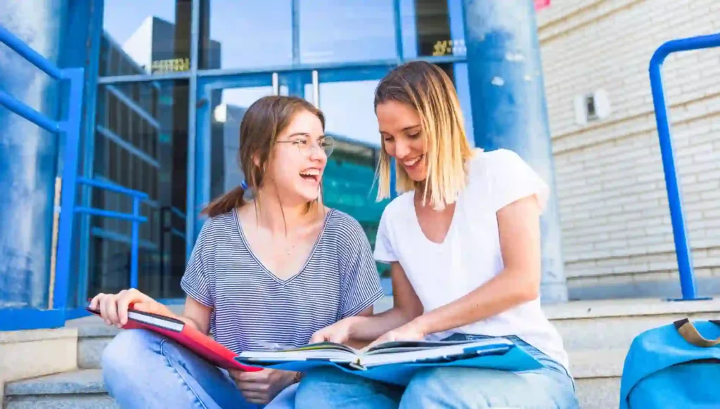 Due ragazze che si sono trasferite in Germania per l'anno all'estero stanno studiando i modi di dire in tedesco seduti sui gradini davanti alla scuola