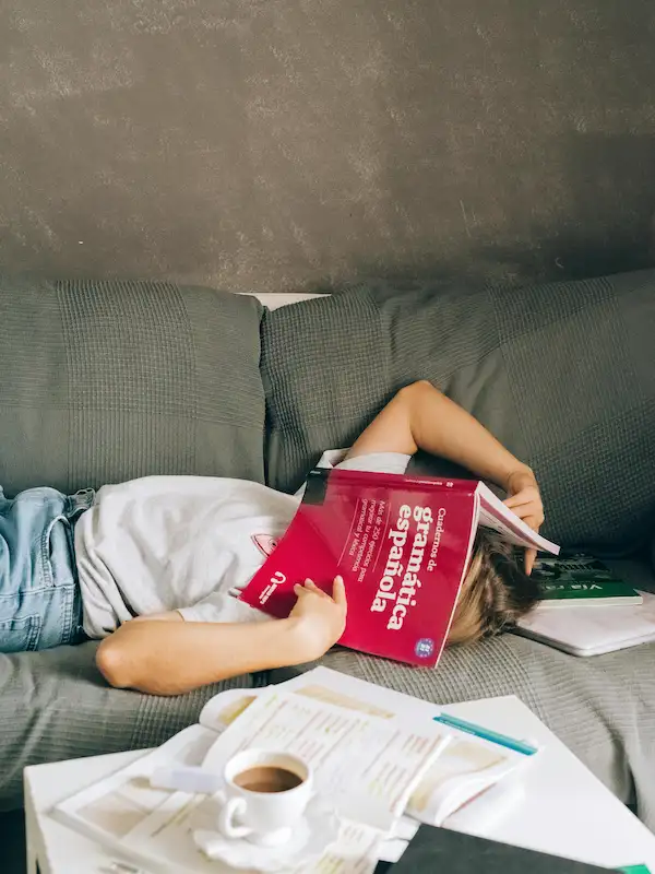 Libri per imparare lo spagnolo: ragazza studia grammatica spagnola distesa su divano grigio con tazza di caffè e quaderni aperti